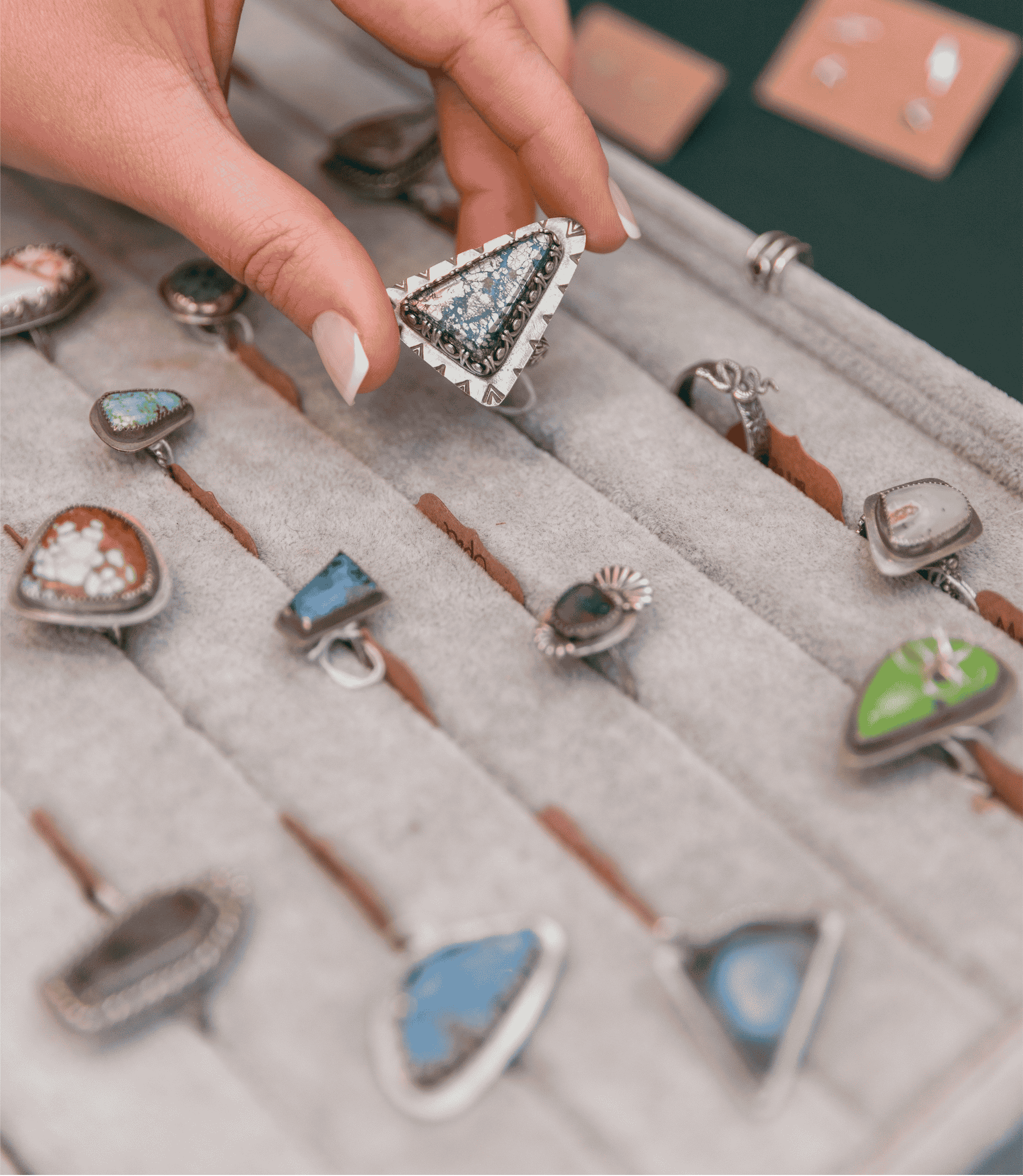 A hand with manicured nails holds a triangular silver ring with a blue and metallic patterned turquoise stone, set against a display tray filled with other rings.